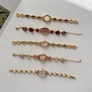 Puede incluir: Cinco relojes de pulsera dorados con diferentes formas de esfera y correas decorativas. Las correas presentan piedras preciosas, perlas y otros adornos. Las esferas son en tonos dorados, rosas y blancos.