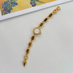 May include: Gold-toned wristwatch with a round, white face and a textured bezel. The band features oval-shaped, brown gemstones and connecting gold links. The watch is a classic accessory.