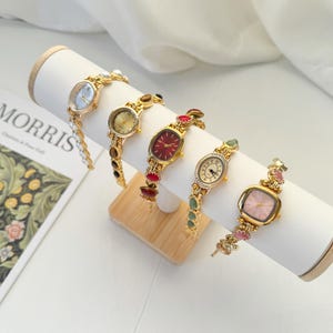 Puede incluir: Cinco relojes de pulsera dorados con varias formas de esfera y correas adornadas con joyas se exhiben en un soporte cil&iacute;ndrico blanco. Las esferas de los relojes incluyen formas ovaladas, redondas y cuadradas, con colores como el rojo, el rosa y el azul claro.