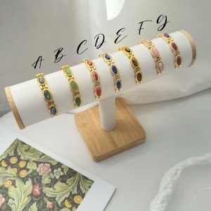K&ouml;nnte beinhalten: Pr&auml;sentation von sieben goldfarbenen Armb&auml;ndern, jedes mit ovalen Edelsteinen in verschiedenen Farben. Die Armb&auml;nder sind auf einem wei&szlig;en, zylindrischen Pr&auml;sentationsst&auml;nder mit Holzsockel angeordnet. Die Edelsteine umfassen blaue, gr&uuml;ne, rote und braune Farbt&ouml;ne.