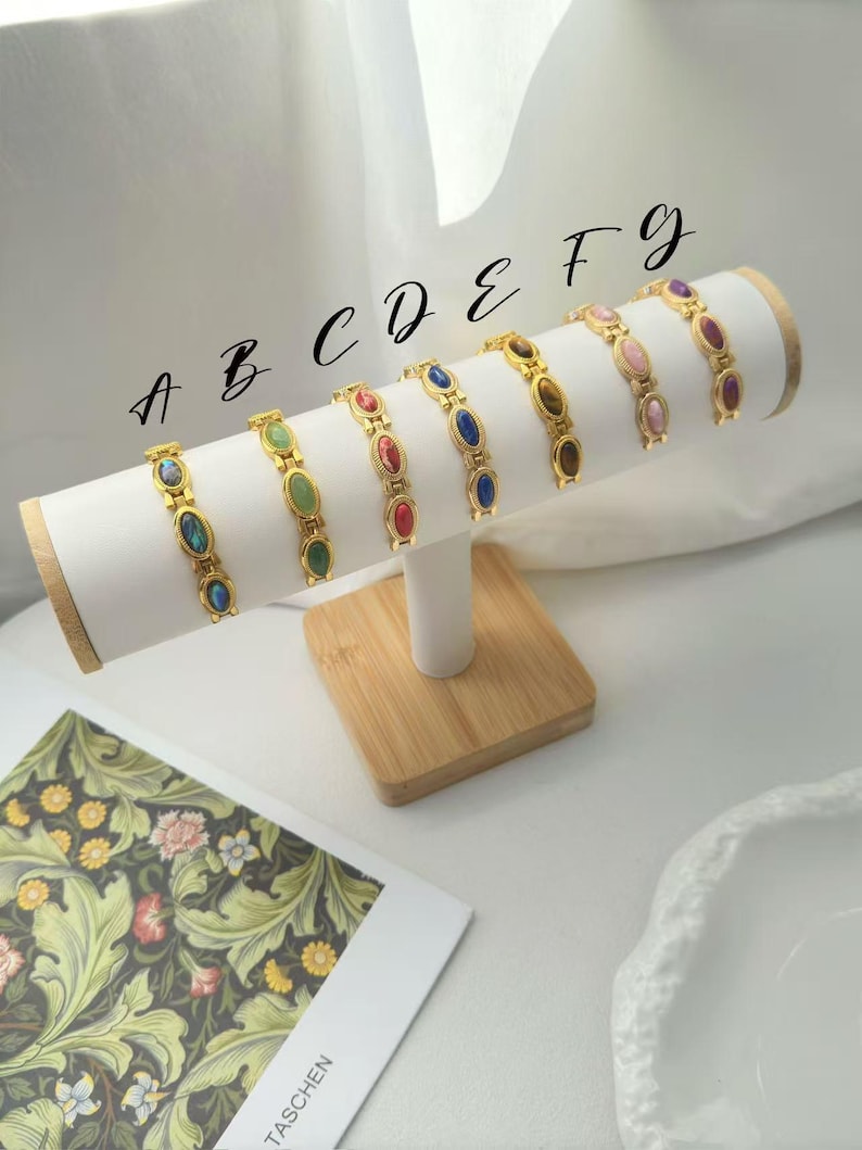 K&ouml;nnte beinhalten: Pr&auml;sentation von sieben goldfarbenen Armb&auml;ndern, jedes mit ovalen Edelsteinen in verschiedenen Farben. Die Armb&auml;nder sind auf einem wei&szlig;en, zylindrischen Pr&auml;sentationsst&auml;nder mit Holzsockel angeordnet. Die Edelsteine umfassen blaue, gr&uuml;ne, rote und braune Farbt&ouml;ne.