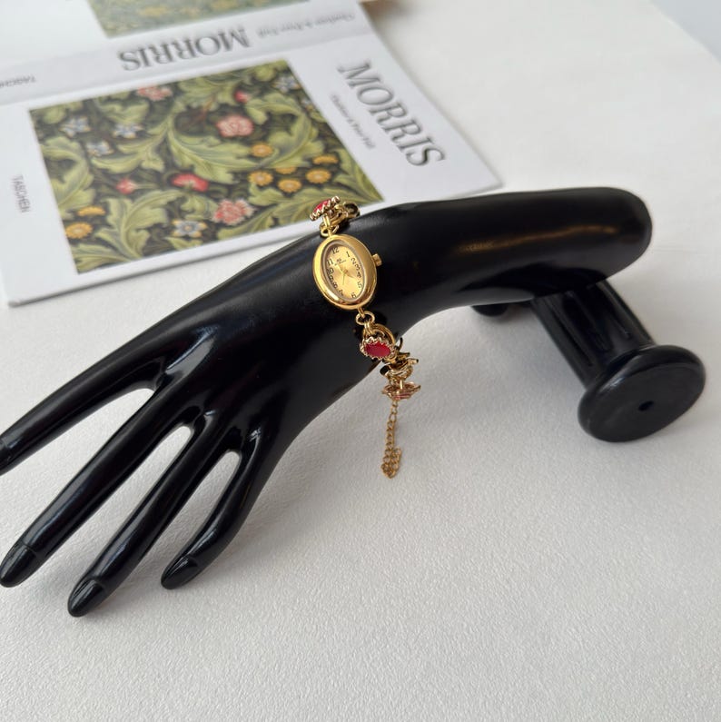 Pu&ograve; includere: Un orologio da polso dorato con un piccolo quadrante ovale e dettagli rossi &egrave; esposto su un espositore nero a forma di mano. L'orologio ha una delicata catena con un ciondolo pendente. Lo sfondo include un libro con la parola "MORRIS".