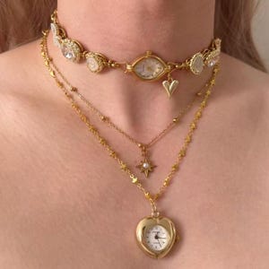 May include: A layered gold-toned necklace set. The top layer is a choker with a watch face and crystal accents. The second layer has a star pendant with a pearl. The bottom layer features a heart-shaped locket watch.
