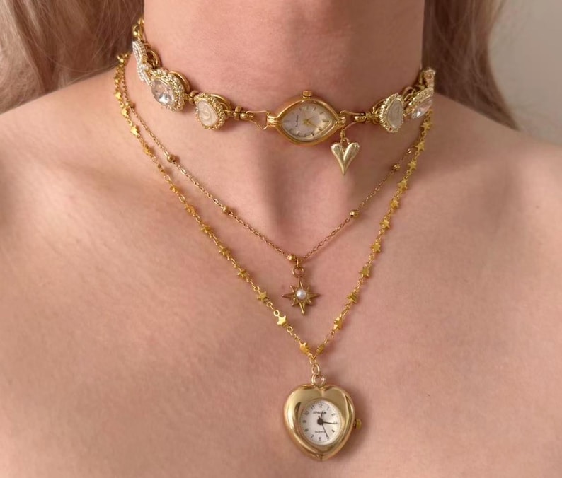 May include: A layered gold-toned necklace set. The top layer is a choker with a watch face and crystal accents. The second layer has a star pendant with a pearl. The bottom layer features a heart-shaped locket watch.