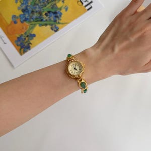 May include: A gold-toned wristwatch with a round face and Roman numeral hour markers. The watch features a textured band and is adorned with green gemstone accents. The watch is on a wrist.
