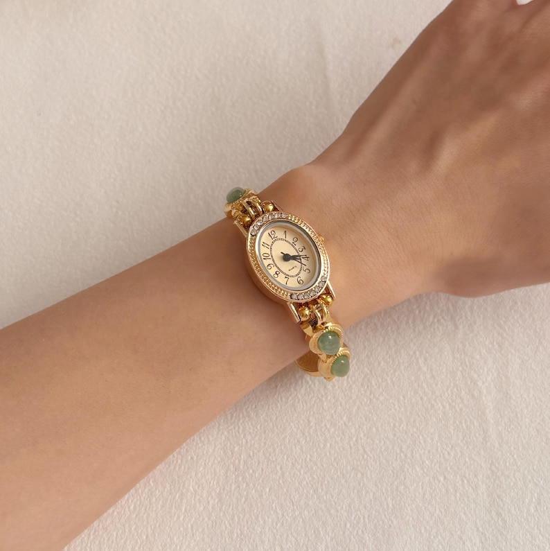 Puede incluir: Un reloj de pulsera dorado con una esfera ovalada y clara. El reloj tiene una correa dorada con detalles verdes en forma de coraz&oacute;n. El reloj se lleva en la mu&ntilde;eca.