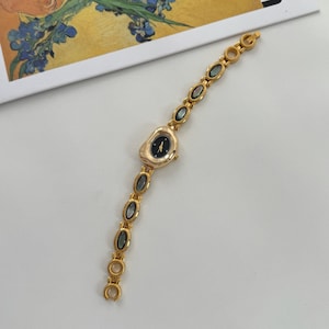May include: A gold-toned wristwatch with a black watch face and a unique, irregular shape. The band features oval-shaped, iridescent accents. The watch is a delicate piece of jewelry, perfect for adding a touch of elegance to any outfit.