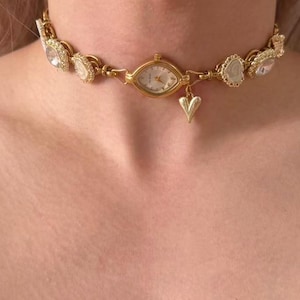 May include: Gold-toned choker necklace featuring a watch face at the center, surrounded by decorative embellishments. A small heart charm dangles below the watch. The necklace is worn around the neck.