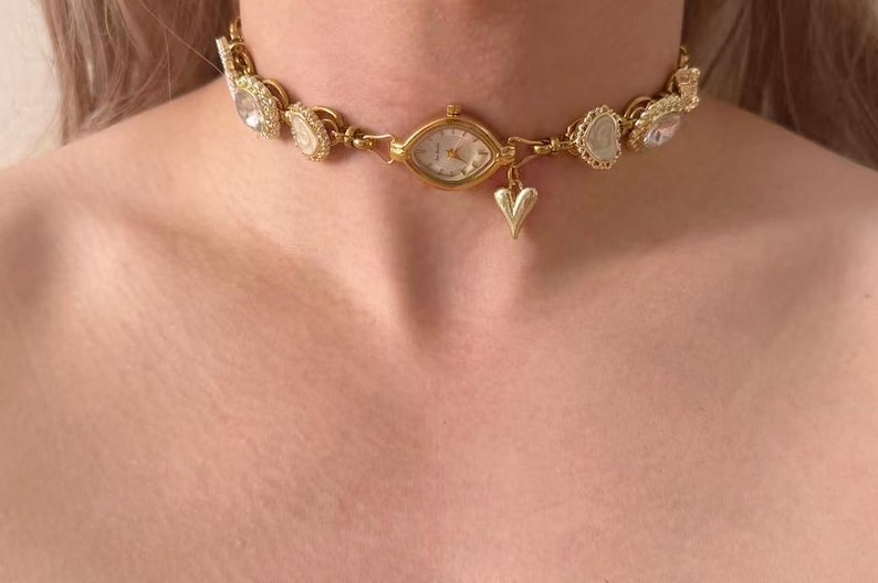 May include: Gold-toned choker necklace featuring a watch face at the center, surrounded by decorative embellishments. A small heart charm dangles below the watch. The necklace is worn around the neck.