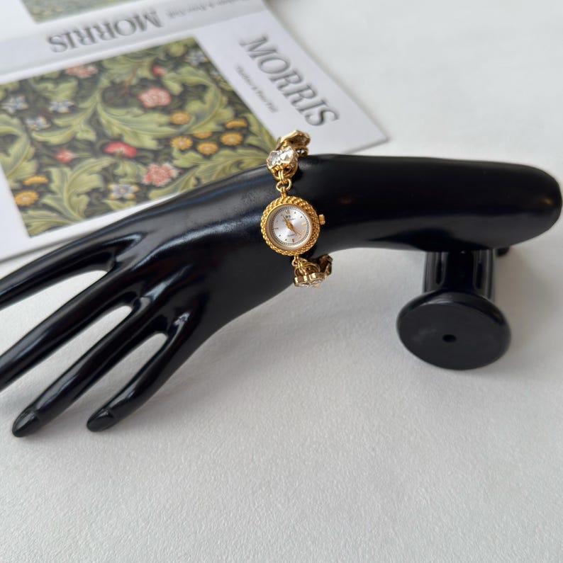 Pu&ograve; includere: Un orologio da polso dorato con quadrante rotondo e bianco. L'orologio &egrave; adornato con cristalli trasparenti ed &egrave; esposto su un supporto nero a forma di mano. L'orologio ha una lunetta intrecciata e un bracciale dorato.