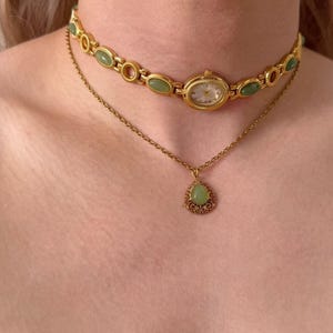 May include: A gold watch choker featuring oval green stones and a small watch face. Beneath, a fine gold chain suspends a teardrop pendant with a green stone and intricate gold filigree. Jewellery.