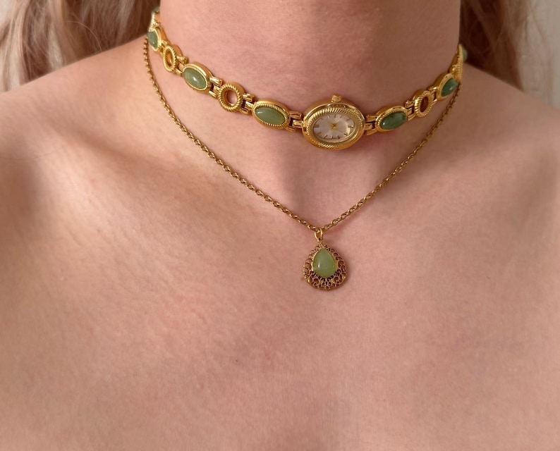 May include: A gold watch choker featuring oval green stones and a small watch face. Beneath, a fine gold chain suspends a teardrop pendant with a green stone and intricate gold filigree. Jewellery.