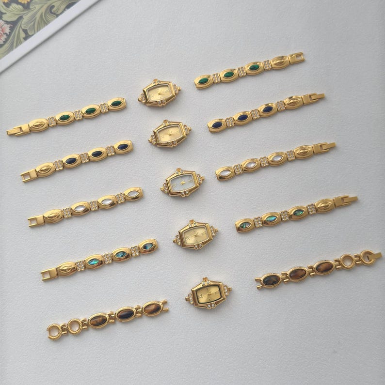 May include: A collection of gold-toned vintage watches and watch bands. The watches have rectangular or oval faces. The bands are adorned with various colored gemstones, including green, blue, and clear crystals. The pieces are arranged on a light-colored surface.