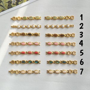 Op de afbeelding: Een verzameling goudkleurige haarspeldjes, elk met een reeks ovale edelstenen in verschillende kleuren, waaronder groen, wit, bruin, roze, rood en abalone. De haarspeldjes zijn in rijen gerangschikt en tonen de verschillende edelsteenopties.