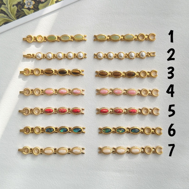 Op de afbeelding: Een verzameling goudkleurige haarspeldjes, elk met een reeks ovale edelstenen in verschillende kleuren, waaronder groen, wit, bruin, roze, rood en abalone. De haarspeldjes zijn in rijen gerangschikt en tonen de verschillende edelsteenopties.