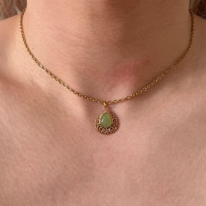 May include: A gold-tone necklace with a fine chain and a teardrop pendant. The pendant has a pale green stone set within an intricate, filigree frame. The necklace is worn around the neck.