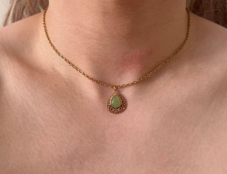 May include: A gold-tone necklace with a fine chain and a teardrop pendant. The pendant has a pale green stone set within an intricate, filigree frame. The necklace is worn around the neck.