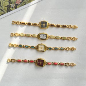 Op de afbeelding: Vier goudkleurige vintage horloges met vierkante of rechthoekige wijzerplaten en decoratieve bandjes. Elk horloge heeft een wijzerplaat in een andere kleur, waaronder zwart, wit en cr&egrave;me. De bandjes zijn versierd met verschillende gekleurde stenen.