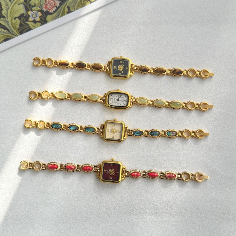 Op de afbeelding: Vier goudkleurige vintage horloges met vierkante of rechthoekige wijzerplaten en decoratieve bandjes. Elk horloge heeft een wijzerplaat in een andere kleur, waaronder zwart, wit en cr&egrave;me. De bandjes zijn versierd met verschillende gekleurde stenen.