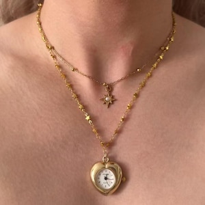 May include: A layered gold necklace featuring a heart-shaped watch pendant and a starburst pendant with a pearl. The necklace has a delicate chain with small star charms. The watch pendant has a white face with black numbers and hands.