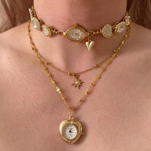 May include: A layered gold-toned necklace set. The top layer is a choker with a watch face and clear stone accents. The second layer has a star pendant, and the bottom layer features a heart-shaped watch pendant.