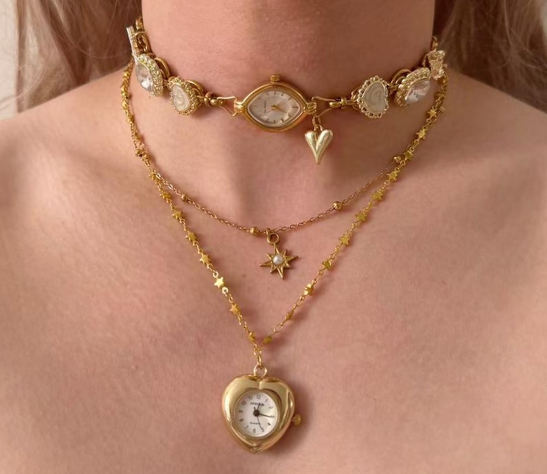 May include: A layered gold-toned necklace set. The top layer is a choker with a watch face and clear stone accents. The second layer has a star pendant, and the bottom layer features a heart-shaped watch pendant.