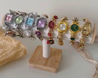 May include: A collection of eight vintage-style wristwatches displayed on a white cylindrical stand with a wooden base. The watches feature square or oval faces in various colors, including pink, green, blue, and gold, with decorative embellishments and charms.