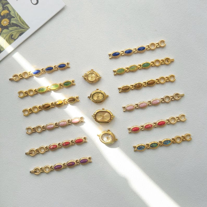 K&ouml;nnte beinhalten: Eine Sammlung goldfarbener Uhrenkomponenten und Armb&auml;nder. Die Armb&auml;nder weisen ovale, farbenfrohe Edelsteine in verschiedenen Farbt&ouml;nen auf, darunter Blau, Gr&uuml;n, Rosa, Rot und Lila. Mehrere Zifferbl&auml;tter sind ebenfalls sichtbar.
