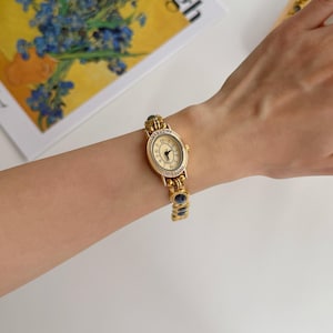 May include: A gold-toned wristwatch with an oval face and a gold band. The watch face has black numerals and hands. The band features blue and clear stones. The watch is on a wrist.