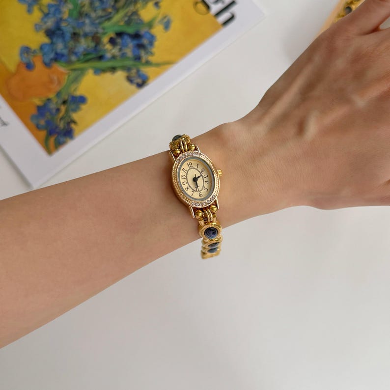 May include: A gold-toned wristwatch with an oval face and a gold band. The watch face has black numerals and hands. The band features blue and clear stones. The watch is on a wrist.