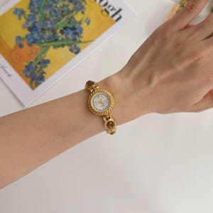 May include: A gold-toned wristwatch with a round face and a textured bezel. The watch has a white dial with gold hands and hour markers. The band features decorative links. The watch is on a wrist.