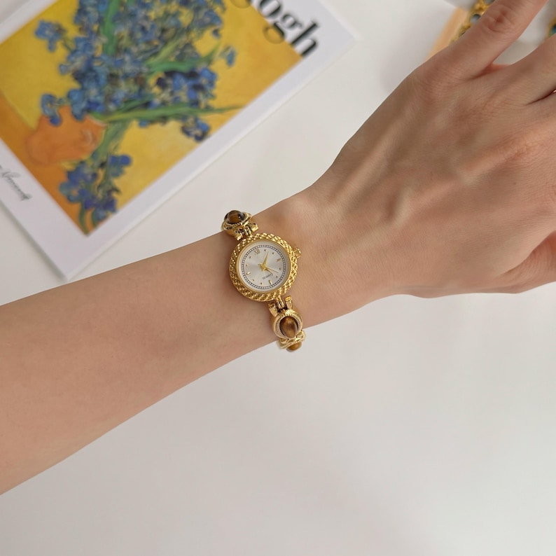 May include: A gold-toned wristwatch with a round face and a textured bezel. The watch has a white dial with gold hands and hour markers. The band features decorative links. The watch is on a wrist.