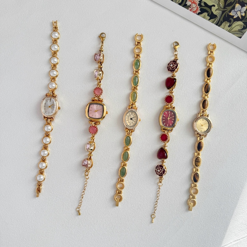 Puede incluir: Cinco relojes de pulsera dorados con diversos elementos decorativos. Los relojes presentan esferas de diferentes colores, incluyendo blanco, rosa y rojo. Las correas est&aacute;n adornadas con perlas, piedras preciosas y detalles florales. Cada reloj tiene un dise&ntilde;o &uacute;nico.