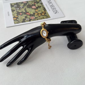 Pu&ograve; includere: Un orologio da polso dorato con quadrante ovale e quadrante bianco. L'orologio &egrave; attaccato a un delicato braccialetto a catena. L'orologio &egrave; esposto su un supporto nero a forma di mano, con un libro sullo sfondo.