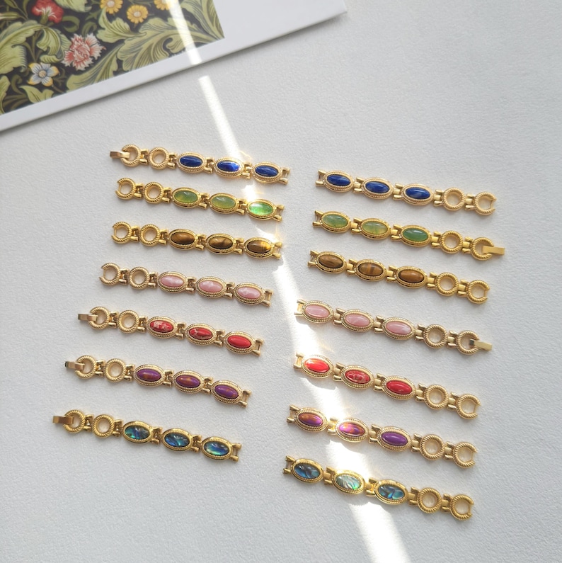 K&ouml;nnte beinhalten: Goldfarbene Armb&auml;nder mit ovalen, farbigen Edelsteinen. Jedes Armband hat eine Reihe von Edelsteinen in verschiedenen Farben, darunter Blau, Gr&uuml;n, Rosa, Rot und Lila. Die Armb&auml;nder sind auf einer wei&szlig;en Oberfl&auml;che angeordnet.