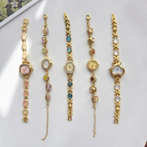 Könnte beinhalten: Fünf goldfarbene Armbanduhren mit dekorativen Gliederarmbändern. Die Uhren verfügen über verschiedene Zifferblattformen wie rund, quadratisch und herzförmig, akzentuiert mit bunten Steinen, irisierenden Einlagen und filigranen Metalldetails.