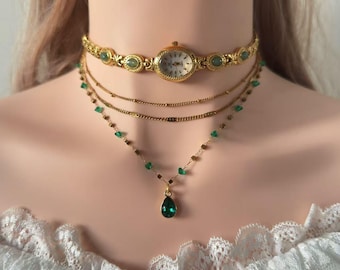 May include: Gold-toned layered necklace set. The top layer is a choker with a watch face and green accents. The second and third layers are delicate chains. The bottom layer features green teardrop pendant.