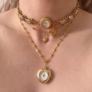 May include: A layered gold-toned necklace featuring a watch pendant and charms. The top layer is a choker with a small watch face. Below, a sun and star charm hang. The bottom layer has a heart-shaped watch pendant.