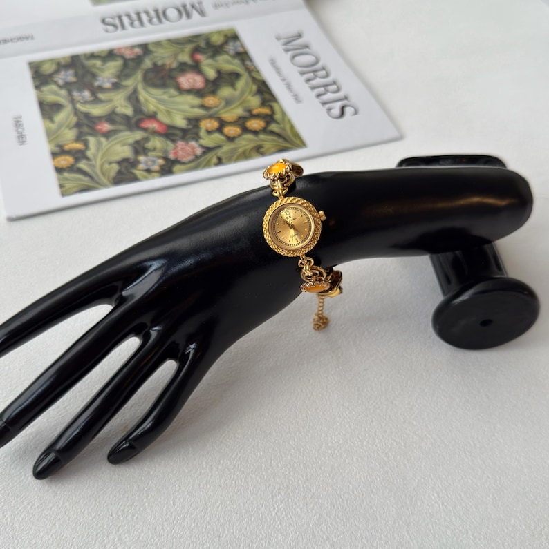 Pu&ograve; includere: Un orologio da polso dorato con quadrante rotondo e catena delicata. L'orologio presenta una gemma gialla nella parte superiore e un ciondolo decorativo appeso in basso. L'orologio &egrave; esposto su un supporto nero a forma di mano.