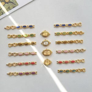 K&ouml;nnte beinhalten: Eine Sammlung goldfarbener Vintage-Uhren und passender Armb&auml;nder. Die Armb&auml;nder sind mit ovalen Edelsteinen in verschiedenen Farben versehen, darunter Blau, Gr&uuml;n, Braun, Rosa, Rot und Lila. Die Uhren haben goldene Geh&auml;use und wei&szlig;e oder goldene Zifferbl&auml;tter.