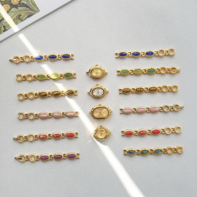 K&ouml;nnte beinhalten: Eine Sammlung goldfarbener Vintage-Uhren und passender Armb&auml;nder. Die Armb&auml;nder sind mit ovalen Edelsteinen in verschiedenen Farben versehen, darunter Blau, Gr&uuml;n, Braun, Rosa, Rot und Lila. Die Uhren haben goldene Geh&auml;use und wei&szlig;e oder goldene Zifferbl&auml;tter.
