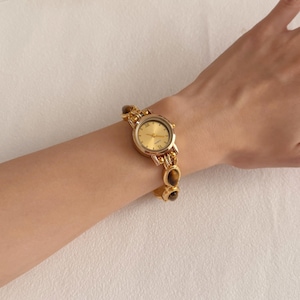 Puede incluir: Reloj de cuarzo dorado con esfera redonda y pulsera a juego. La esfera del reloj es dorada con manecillas doradas y n&uacute;meros romanos. La pulsera presenta piedras ovaladas marrones y eslabones dorados.