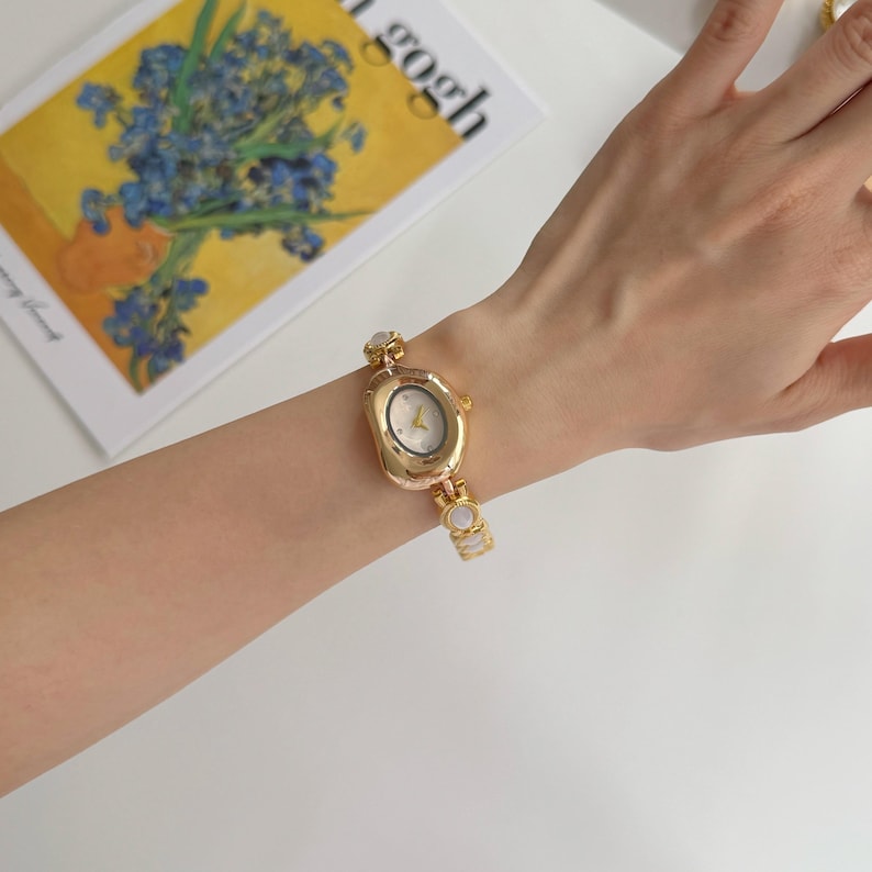 May include: Gold-toned wristwatch with an oval face and a delicate bracelet band. The watch features a white watch face with gold hands and markers. The band is adorned with small, round, white accents. The watch is displayed on a person's arm.