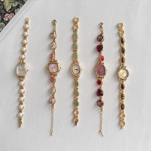 Puede incluir: Cinco relojes de pulsera dorados con varios diseños. Un reloj presenta perlas, otro piedras preciosas rosas, un tercero piedras verdes, un cuarto corazones rojos y el último piedras marrones. Cada reloj tiene un diseño de esfera diferente.