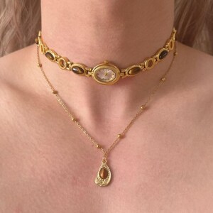 May include: A gold watch choker and a delicate gold necklace with a pendant. The watch choker features an oval watch face and brown stone accents. The necklace has a teardrop-shaped pendant with a brown stone.