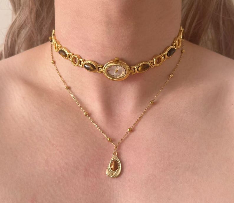 May include: A gold watch choker and a delicate gold necklace with a pendant. The watch choker features an oval watch face and brown stone accents. The necklace has a teardrop-shaped pendant with a brown stone.