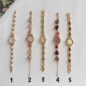 Puede incluir: Cinco relojes de pulsera dorados con varios elementos decorativos. Los relojes tienen diferentes esferas y correas de colores adornadas con perlas, piedras preciosas y dijes en forma de coraz&oacute;n. Cada reloj tiene un dise&ntilde;o &uacute;nico, con los n&uacute;meros del 1 al 5 debajo.