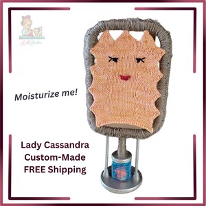 Peut inclure: Une pièce décorative Lady Cassandra faite sur mesure. La pièce présente un visage tricoté de couleur pêche avec des yeux noirs et une bouche rouge, le tout dans un cadre gris. Le texte "Moisturize me!" est également visible.
