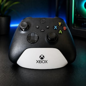 Könnte beinhalten: Ein schwarzer kabelloser Videospiel-Controller mit einem weißen Sockelbereich, der das schwarze Xbox-Logo und den Schriftzug trägt. Der Controller verfügt über zwei schwarze Analogsticks, ein Steuerkreuz und vier farbige Aktionstasten Y, X, B und A.