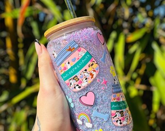 May include: A clear glass tumbler with a bamboo lid and a glass straw. The tumbler is filled with purple glitter and decorated with dog-themed stickers, including the text "ANTI DEPRESSANTS", hearts, a rainbow, and a paw print.